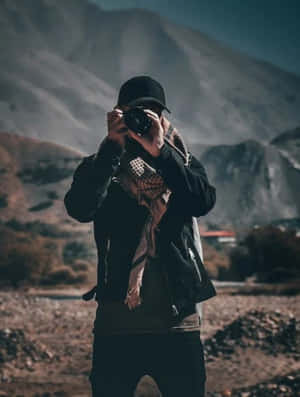喺山區地形中，一個人穿著黑色外套同帽，手持相機。