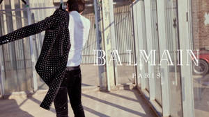 身穿圓點西裝外套的男子在陽光明媚的城市小巷中走遠，帶有Balmain Paris文字。