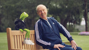 一位身穿藍色運動服的長者與穿著連帽衫的青蛙柯密特坐在長椅上。 — 網球 壁紙
