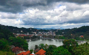 多雲的天空下，一個湖邊小鎮被綠色山丘環繞的風景如畫的景色。 — 湖景 壁紙