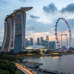 濱海灣金沙和新加坡摩天觀景輪在黃昏時亮起，城市燈光倒映在水面上 — 建築 壁紙