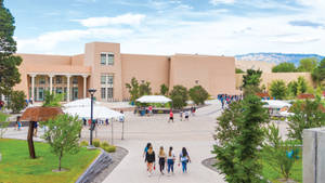 學生在校園小徑上走向一棟米色建築，周圍有綠樹和白色帳篷。