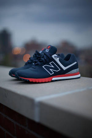 海軍藍色 New Balance 運動鞋（紅色鞋底）在黃昏時分放在城市屋頂邊緣上。 — 鞋子 壁紙