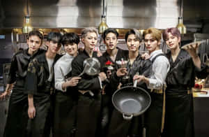 七位K-pop偶像打扮成廚師，在攝影棚廚房裡拿著廚房工具擺姿勢。