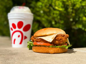脆皮雞肉三文治配芝士、番茄和生菜，旁邊放著Chick-fil-A飲料杯，戶外場景。