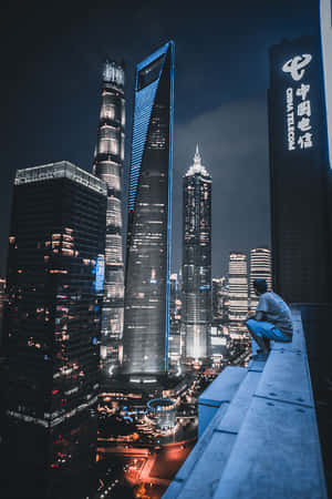 人坐在屋頂邊緣凝視著夜晚燈火通明的摩天大樓 — 天際線 壁紙