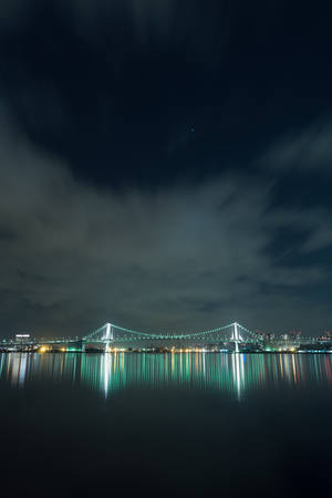 夜晚一座燈火通明的橋樑，其倒影映在雲天下的水面上。