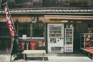 兩部 Kirin 自動售賣機矗立在日本商店外，旁邊有木長椅和紅色橫幅。 — 日本 壁紙