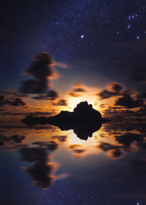 繁星點點的夜空，發光的雲朵倒映在平靜的水面上，岩石島嶼的剪影之上。