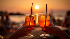 兩隻手在水邊夕陽下用冰凍橙汁飲料碰杯 — 夏日 壁紙