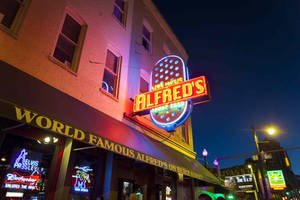霓虹燈照亮的 Alfred's 餐廳和貓王紀念品，位於夜晚的城市街道上。 — 夜晚 GIF 壁紙