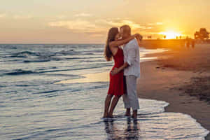 一對情侶在日落時分的沙灘上親吻，海浪圍繞他們的腳。 — 情侶 GIF 壁紙