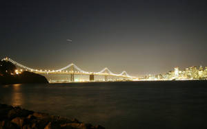 星空下，夜間城市燈光映照水面上的一座亮燈吊橋。
