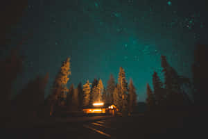 一間溫暖照明的小屋，被松樹環繞，上方是星空。 — 夜空 壁紙