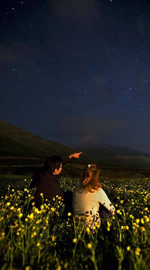 兩個人坐在黃色花田裡，指著山上的星星。