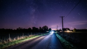 一輛汽車在星空下的夜間行駛在鄉間小路上，可見房屋和圍欄。