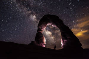 夜晚，一個人站在天然石拱門下，星空下可見銀河。 — 太空 壁紙