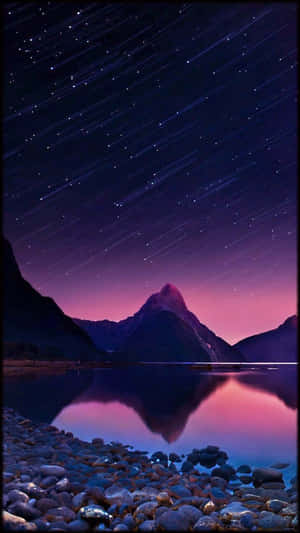 在繁星點點的紫色夜空下，山峰倒映在湖中，湖邊有岩石岸。 — 月色夜空 壁紙