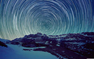 在深藍色夜空下，覆蓋著雪的山脈上方螺旋狀的星軌。 — 星星 壁紙