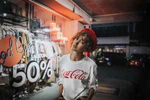 年輕女子戴紅色貝雷帽、身穿可口可樂襯衫，夜晚靠近發光的商店櫥窗。