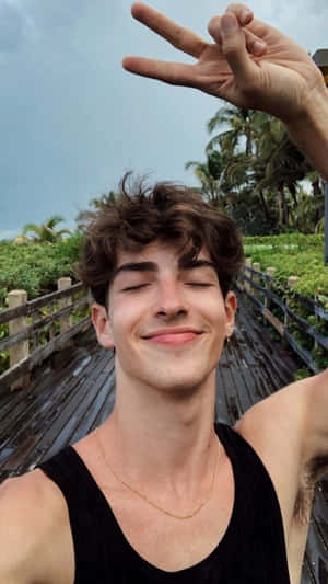 年輕男子閉眼微笑比出和平手勢，站在木橋上，背景有棕櫚樹