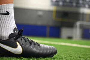 黑色Nike足球鞋，金色swoosh標誌，置於綠色人造草地上。