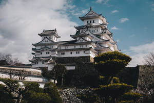 白色日本城堡在石山上，周圍有綠樹和部分多雲的天空