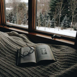 打開的書本連同眼鏡放在有紋理的毯子上，靠近可俯瞰雪覆蓋森林的窗戶。 — 冬日美學 壁紙