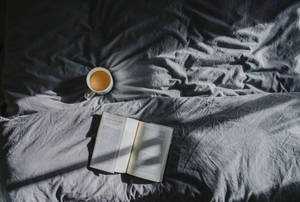 一杯茶和一本打開的書放在灰色床單上，在柔和的晨光中。