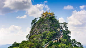 金色寺廟坐落在岩石山頂上，周圍環繞著綠樹和藍天。