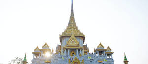 金色泰國寺廟尖塔，中央塔樓後方有陽光四射，映襯著明亮的天空。