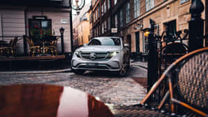 銀色 Mercedes-Benz EQC SUV 停泊在鵝卵石街道上，靠近戶外咖啡座位。 — 城市 壁紙