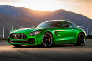 綠色 Mercedes-AMG GT 跑車停在瀝青路上，背景有山脈和風力發電機，夕陽西下。