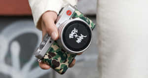 手拿著帶有迷彩機殼和塗鴉鏡頭蓋的銀色 Leica 相機。