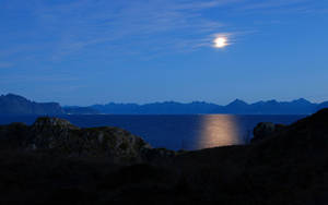 月光在夜晚照亮黑暗岩石海岸和遠處山脈之間的平靜海水。