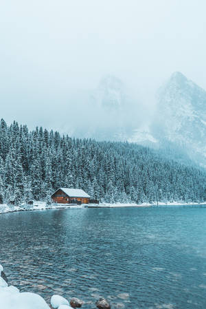冬季藍色湖邊的雪中小屋，有松樹和霧山。