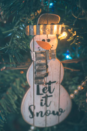 木製雪人裝飾品，帶有「Let it Snow」文字，掛在點亮的聖誕樹上。