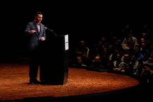 男子在木製舞台上發表演講，觀眾在陰影中。