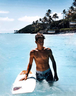 年輕衝浪者在碧綠海水中手持衝浪板，靠近棕櫚樹