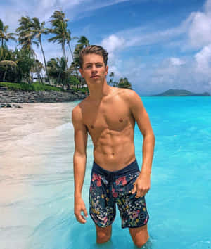年輕男子穿著花卉泳褲站在熱帶海灘的碧綠海水中。