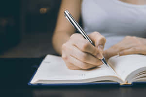 在深色書桌上用銀色筆在筆記本上書寫的手。 — MacBook Pro 14 壁紙