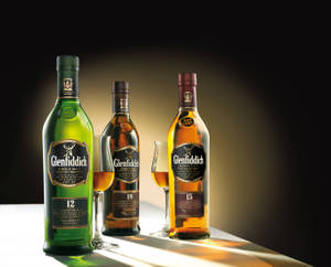 三瓶 Glenfiddich 威士忌酒瓶連酒杯放在發光表面上，背景黑暗。