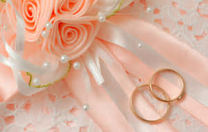 兩枚金色結婚戒指，旁邊有蜜桃色布料玫瑰和帶珍珠的蕾絲。