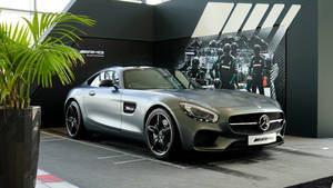 銀色 Mercedes-AMG GT 跑車在展廳內，配有賽車牆面藝術和一棵盆栽棕櫚樹。