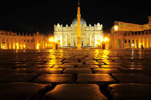 聖伯多祿大教堂夜景，金色燈光映照在濕漉漉的鵝卵石上。