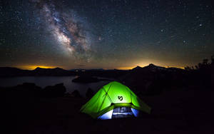 在星空下夜間搭建的發光綠色帳篷，湖泊和山脈上方可見銀河。