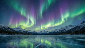 綠色和紫色極光反射在冰凍的湖泊上，周圍是雪山，星空璀璨