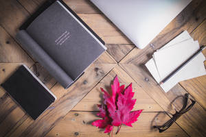 木製書桌上有手提電腦、筆記本、眼鏡和一片紅楓葉。