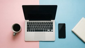 銀色手提電腦、咖啡杯、電話和筆記本在粉紅色和藍色背景上。
