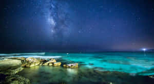 繁星點點的夜空，銀河系在碧綠色的海洋和岩石海岸上方。 — 藍天 壁紙